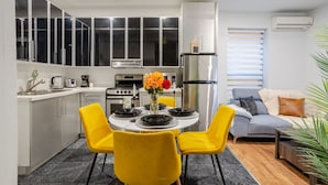 A modern kitchen with white cabinets, a stainless steel refrigerator, and a dining area with yellow chairs and a round table. - Charming 1BDR – Prime Location on Bishop St (Montréal)