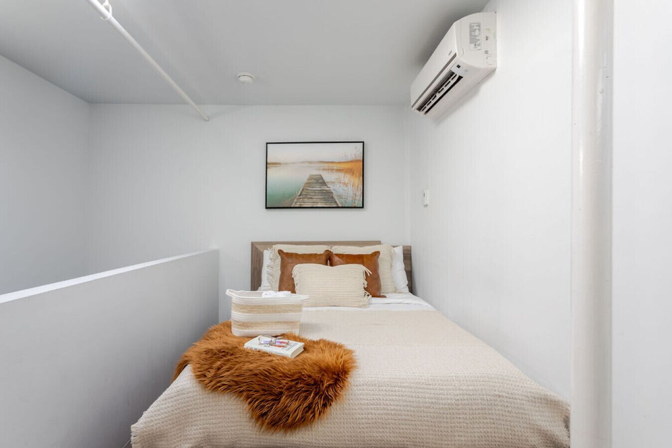 Quarto de hotel estreito com uma cama, um quadro pendurado na parede e um aparelho de ar condicionado.