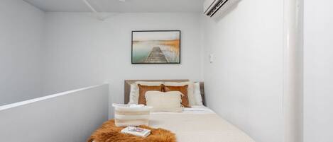 Quarto de hotel estreito com uma cama, um quadro pendurado na parede e um aparelho de ar condicionado.