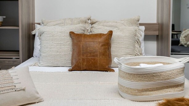 Ein Bett mit gestreifter Kissen, einem braunen Lederkissen und einem gewebten Korb auf einem weißen Teppich.
