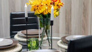 Ein Esstisch mit Glasplatte, umgeben von gewebten Platzsets, Tellern und einer Vase mit gelben Blumen.