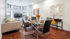 A modern living room with a glass dining table, black leather chairs, a sofa, and a TV. - Vibrant MTL Living – 1BDR in a Top Location (Montréal)