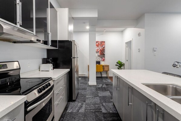 A modern kitchen with stainless steel appliances, a central island, and a view into the dining area. - Vibrant MTL Living – 1BDR in a Top Location (Montréal)
