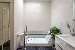 A modern bathroom with a glass-enclosed bathtub, a wooden shelf, a potted plant, and a white sink. - Vibrant MTL Living – 1BDR in a Top Location (Montréal)
