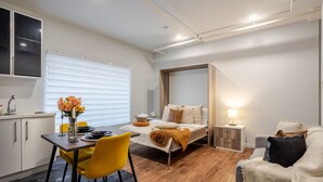 A modern bedroom with a bed, bedside table, and a nightstand. There is a window with blinds, a desk with a chair, and a sofa. - Spacious Bishop Apartment –Prime Downtown Stay (Montréal)