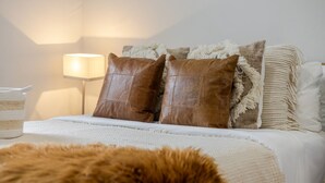 Una cama bien hecha con una colcha blanca y dos almohadas de cuero marrón, un plaid blanco mullido y una lámpara en una mesita de noche.