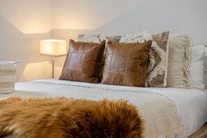 Un lit bien fait, avec une couette blanche et deux coussins en cuir marron, un plaid blanc moelleux et une lampe sur une table de chevet.
