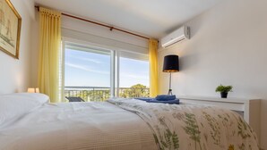A bedroom with a large bed, a window with yellow curtains, a black floor lamp, and a small potted plant on a bedside table.