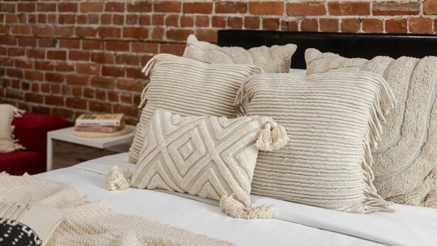 A bed with multiple pillows and a blanket, a brick wall, and a small table with a book.