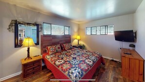 A bedroom with a wooden headboard, a large bed with a colorful quilt, two bedside lamps, a mirror, a television mounted on the wall, and a small potted plant.