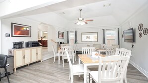 A spacious living area with a dining table, white chairs, a kitchen area with cabinets, a ceiling fan, and a TV mounted on the wall. - 8BR Home • Near IAH & Downtown • Sleeps 17 • Spacious & Private backyard for BBQ (Humble)