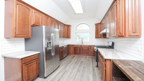 A modern kitchen with wooden cabinets, a stainless steel refrigerator, a built-in oven, and a window with blinds. - 8BR Home • Near IAH & Downtown • Sleeps 17 • Spacious & Private backyard for BBQ (Humble)
