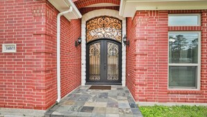 A red brick house with a black double door, a small porch, and a window with blinds. - 8BR Home • Near IAH & Downtown • Sleeps 17 • Spacious & Private backyard for BBQ (Humble)