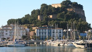 Un puerto deportivo con barcos amarrados, un edificio con varias ventanas y una colina con una fortificación al fondo.