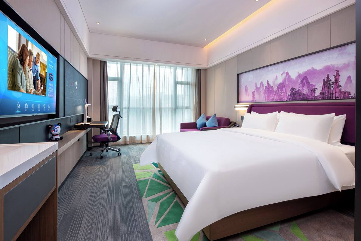Une chambre d’hôtel moderne dotée d’un grand lit, d’un bureau avec un ordinateur, d’une télévision et d’une grande fenêtre avec des rideaux.