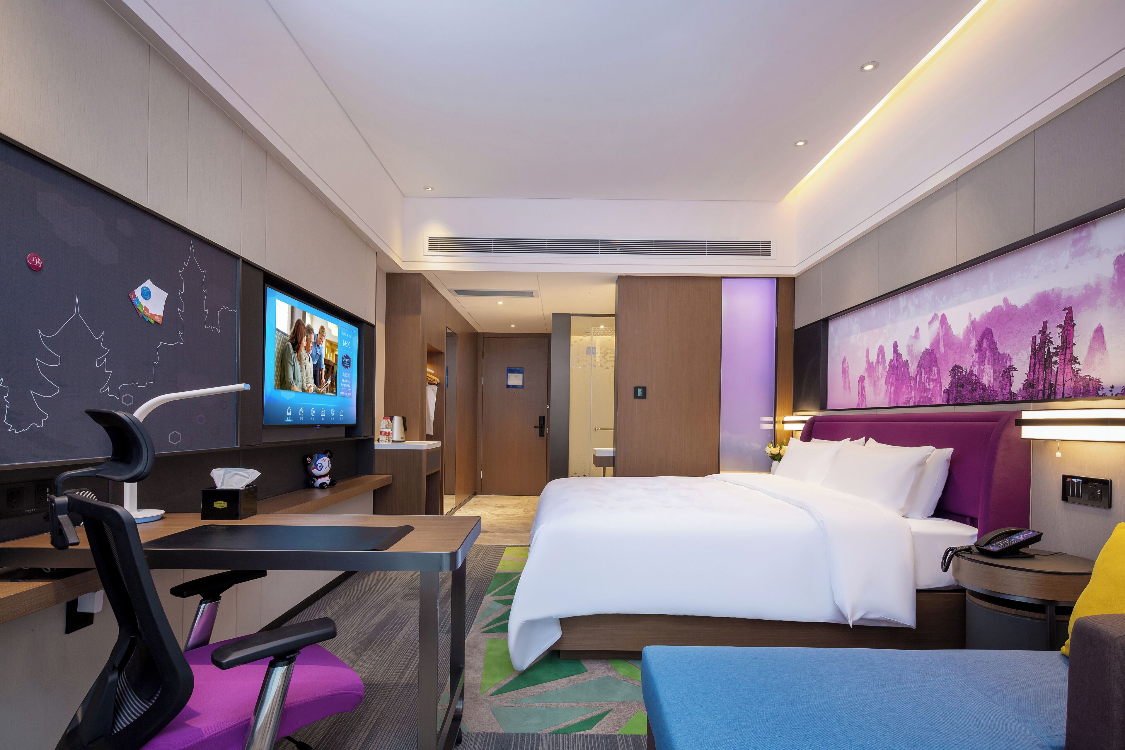 Een moderne hotelkamer met een bed, bureau, stoel en een grote tv aan de wand.