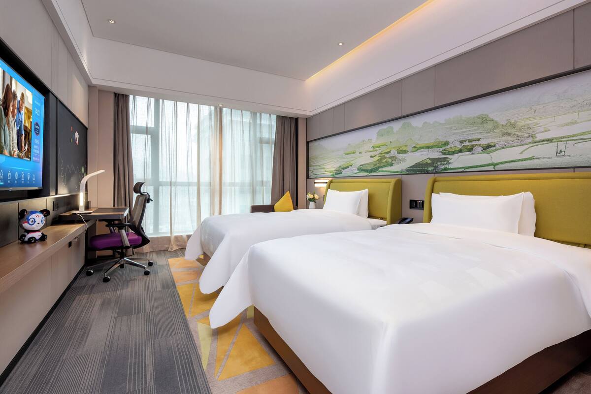 Une chambre d’hôtel avec deux lits, un bureau avec un ordinateur, une télévision et une grande fenêtre avec des rideaux.