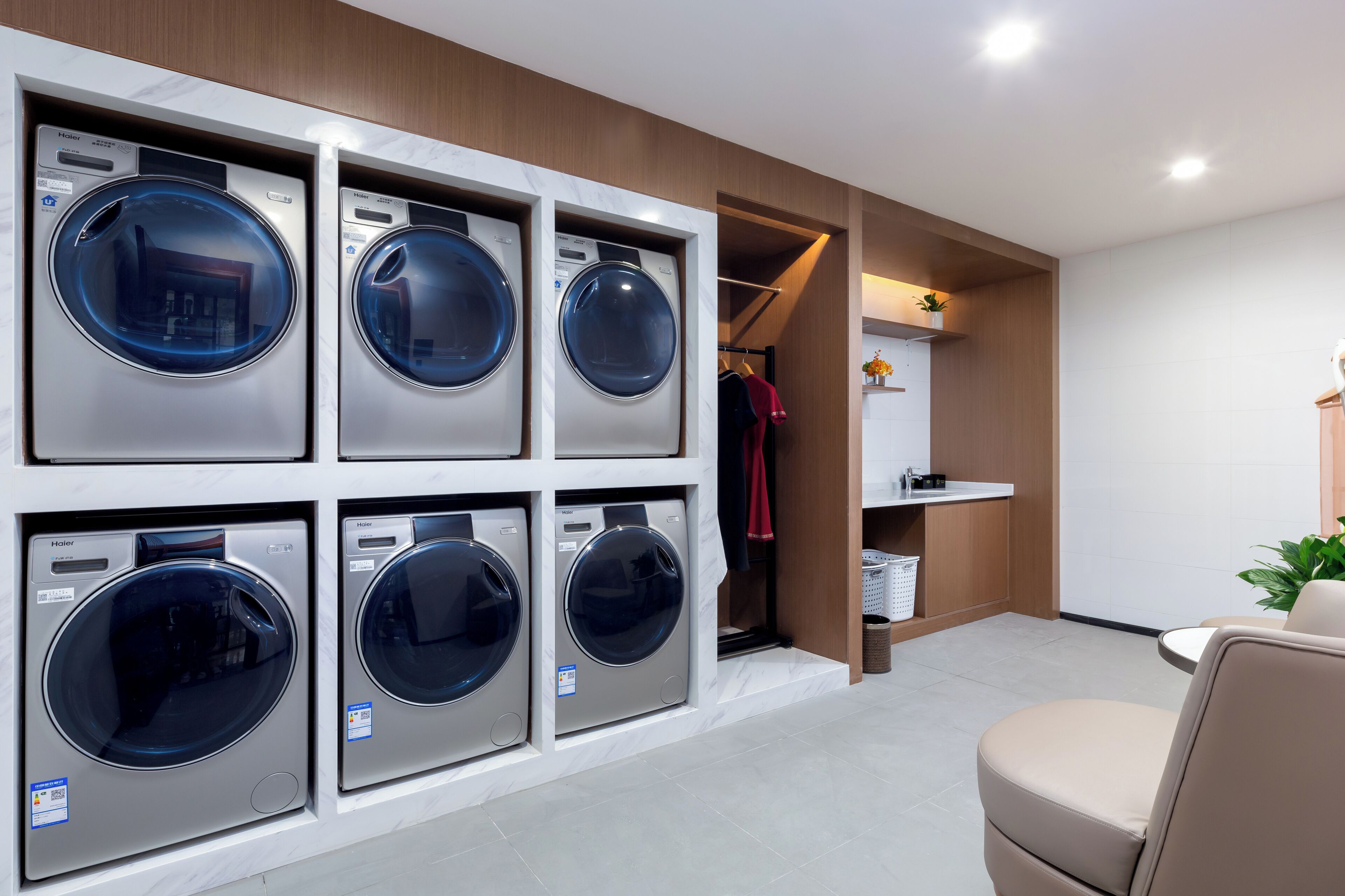 A laundry room with six front-loading washers and dryers, a wooden closet, and a seating area with a chair.