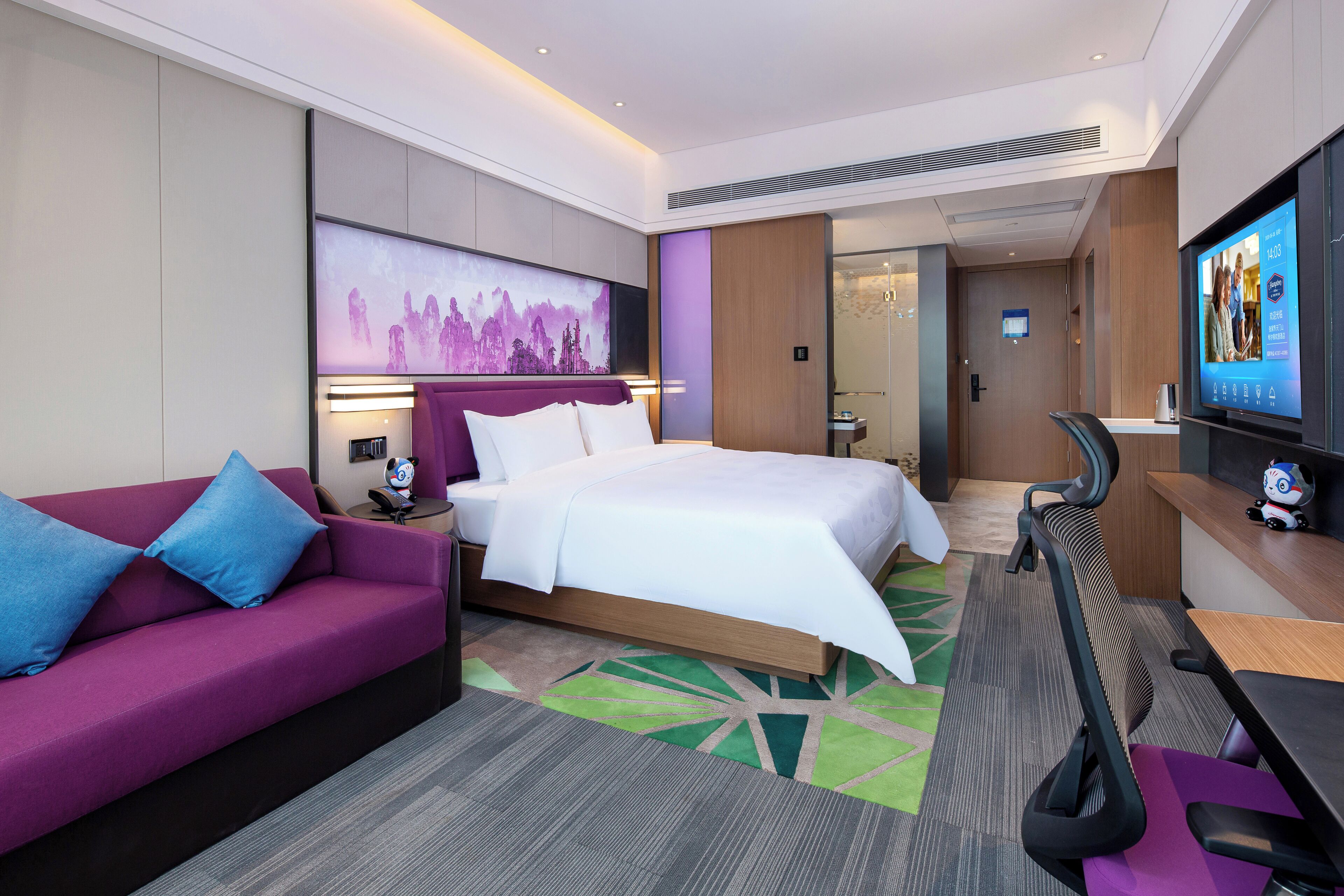 A modern hotel room with a bed, a purple sofa, a desk with a chair, and a TV on the wall.