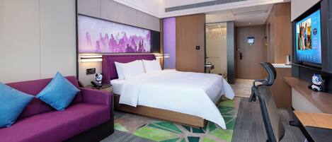 Una habitación de hotel moderna con una cama, un sofá morado, un escritorio con una silla y un televisor en la pared.