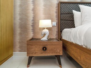 Une table de chevet en bois, sur laquelle sont posés un objet décoratif blanc et une lampe, se trouve à côté d’un lit doté d’une tête de lit.