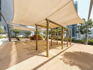 A playground with a climbing structure and swings, surrounded by a sandy area and trees.