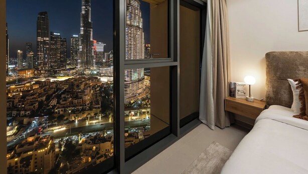 Habitación de hotel con un ventanal grande que ofrece vistas nocturnas de la ciudad, una cama con cabecero, una mesita de noche con lámpara y un cuadro enmarcado en la pared.