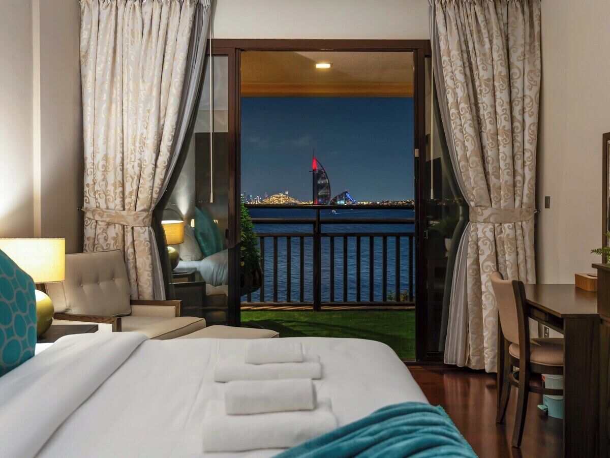 Une chambre d’hôtel avec un lit, un bureau, une chaise et une vue sur une étendue d’eau, avec un bâtiment visible au loin.