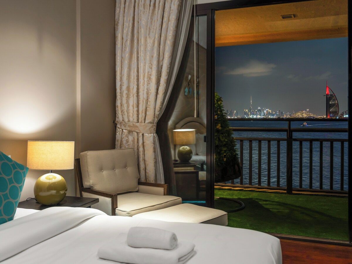 Een hotelkamer met een bed, een stoel, een lamp en uitzicht op een stadshemel en water.