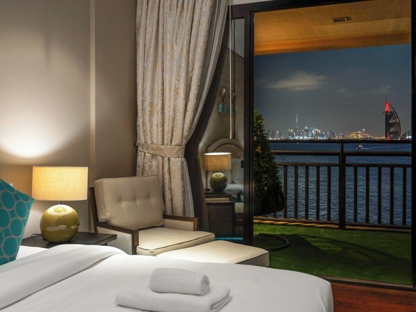 A hotel room with a bed, a chair, a lamp, and a view of a city skyline and water.