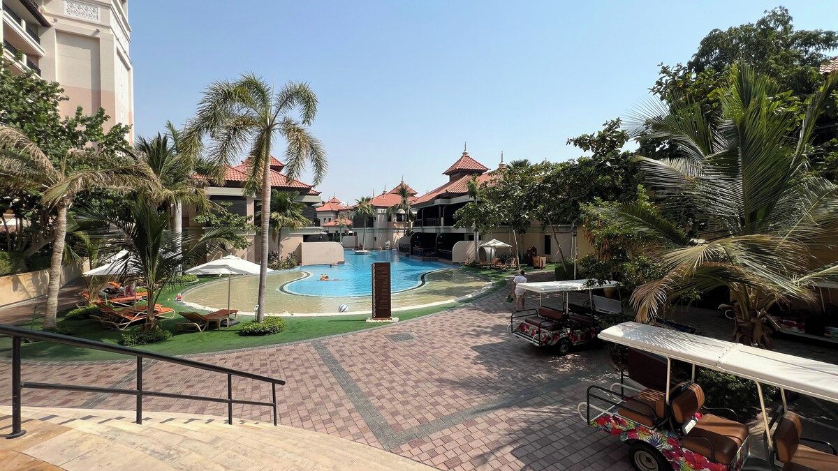 Un espace piscine aménagé dans un complexe hôtelier, avec des palmiers, des chaises longues et un coin salon abrité.