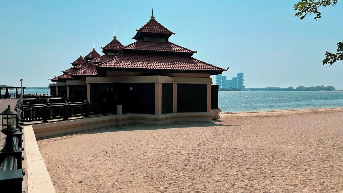 Un pavillon traditionnel à plusieurs niveaux, surmonté d’un toit de tuiles, situé sur une plage de sable, avec une étendue d’eau en arrière-plan.