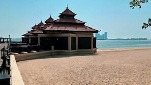 A traditional pavilion with multiple tiers and a tiled roof, situated on a sandy beach with a body of water in the background.