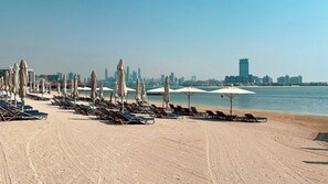 Uma praia com fileiras de espreguiçadeiras e guarda-sóis, céu claro e o skyline de uma cidade ao fundo.