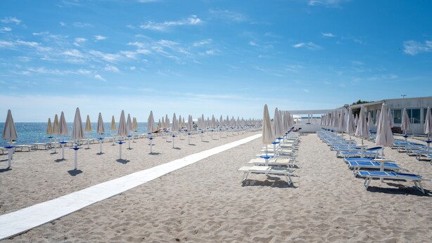 Une plage bordée de rangées de chaises longues et de parasols, un chemin blanc et un bâtiment visible au loin.