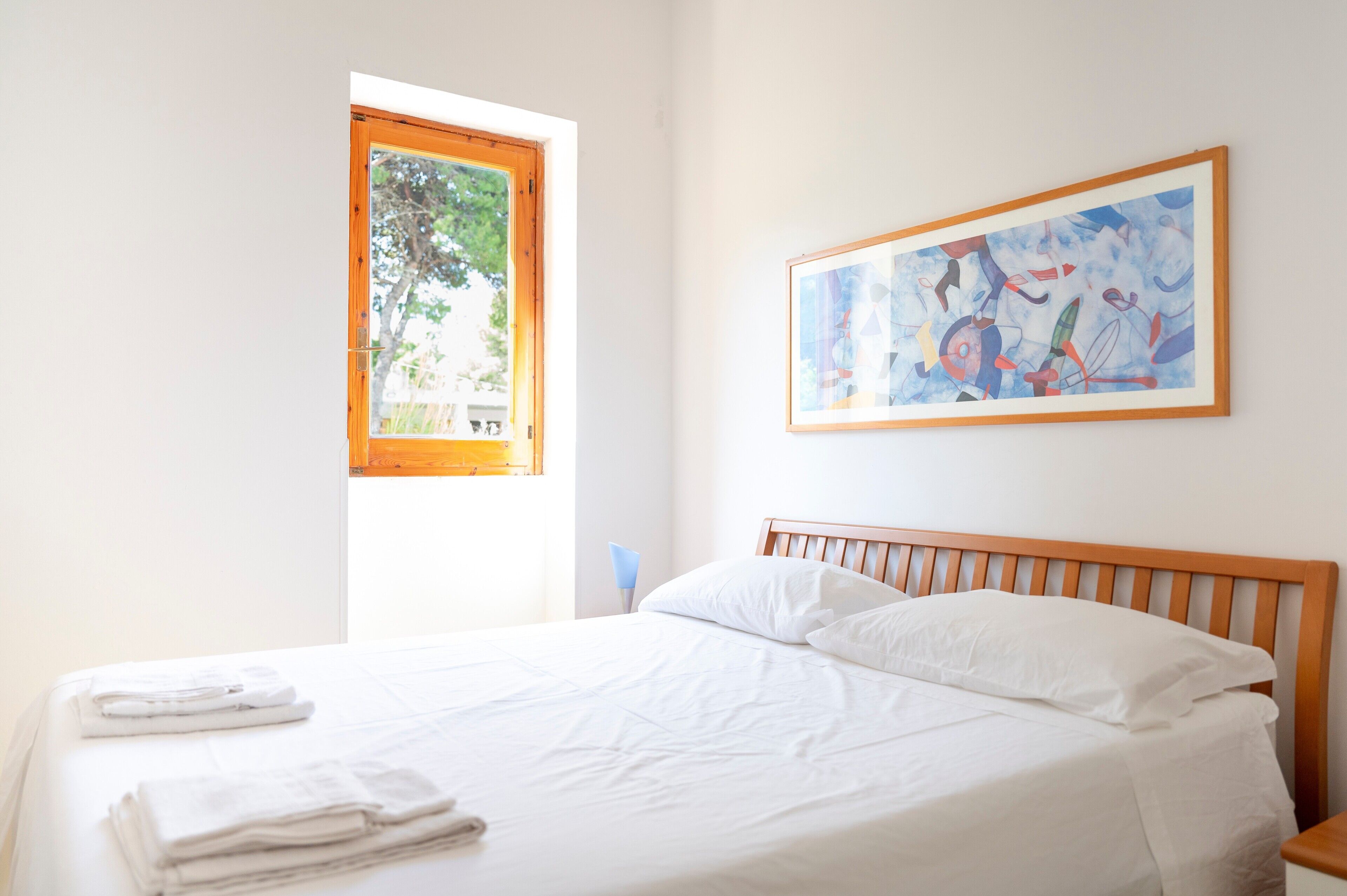 Un letto rifatto con lenzuola bianche, una finestra con vista sul verde e un quadro astratto appeso al muro.