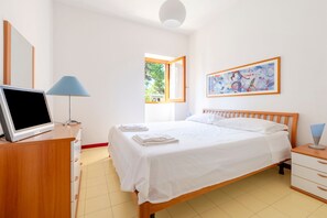 Een slaapkamer met een bed, een bureau met een computer, een lamp, een schilderij en een raam.