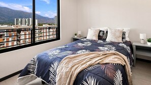 A hotel room with a bed, a nightstand, a window with a view of buildings and mountains, and a view of the sky. - Oceana Suites La Florida Friolan Roa (Santiago)