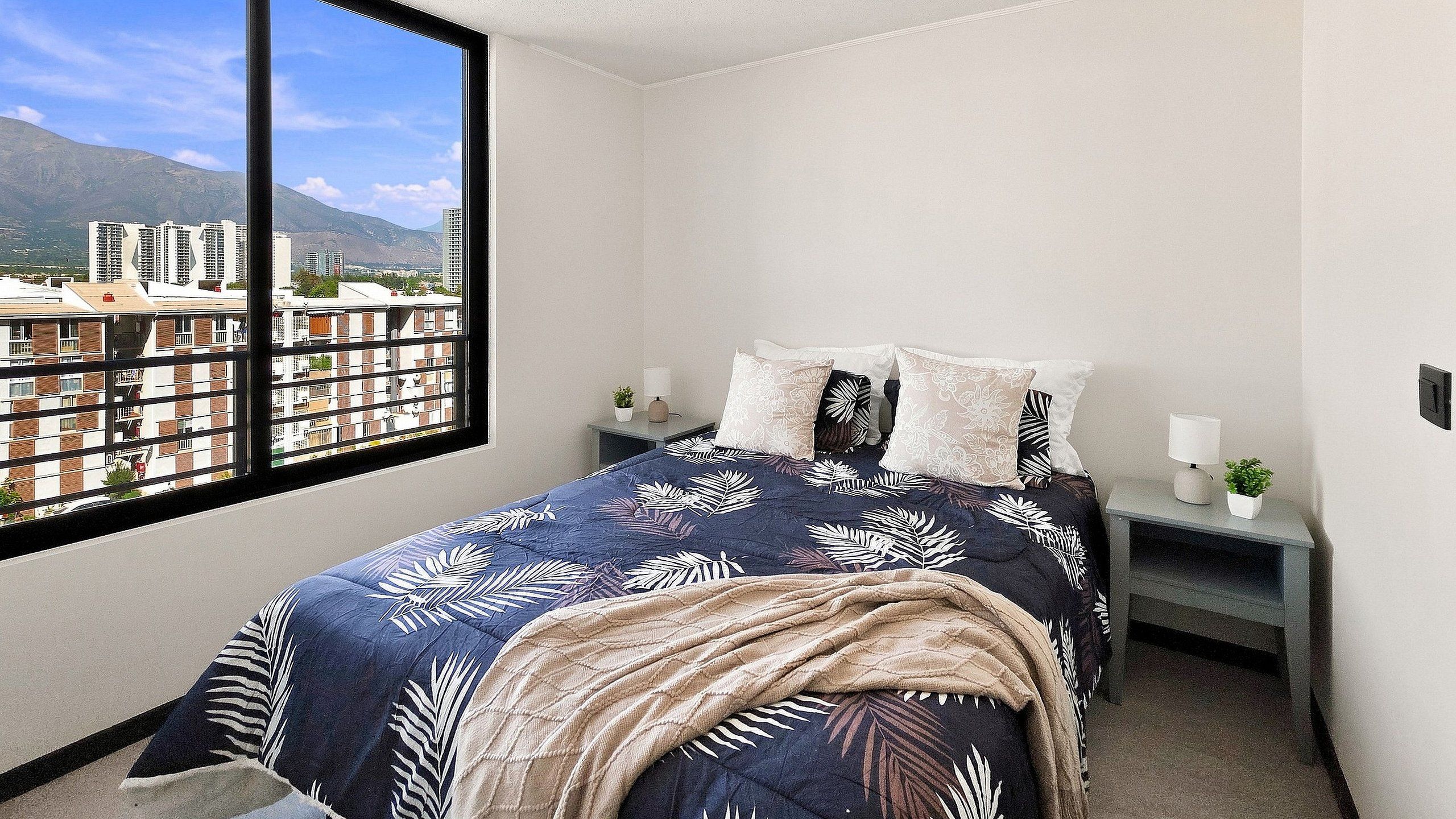 Een slaapkamer met een groot raam, een bed met beddengoed met een patroon, een nachtkastje met een lamp, en een uitzicht op gebouwen en bergen.