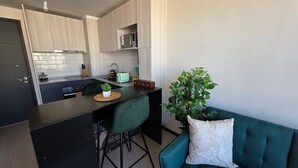 Kompakti keittiö, jossa on keskeinen saareke, ruokailutila, jossa on vihreä tuoli, sekä turkoosi sohva koristetyynyllä.