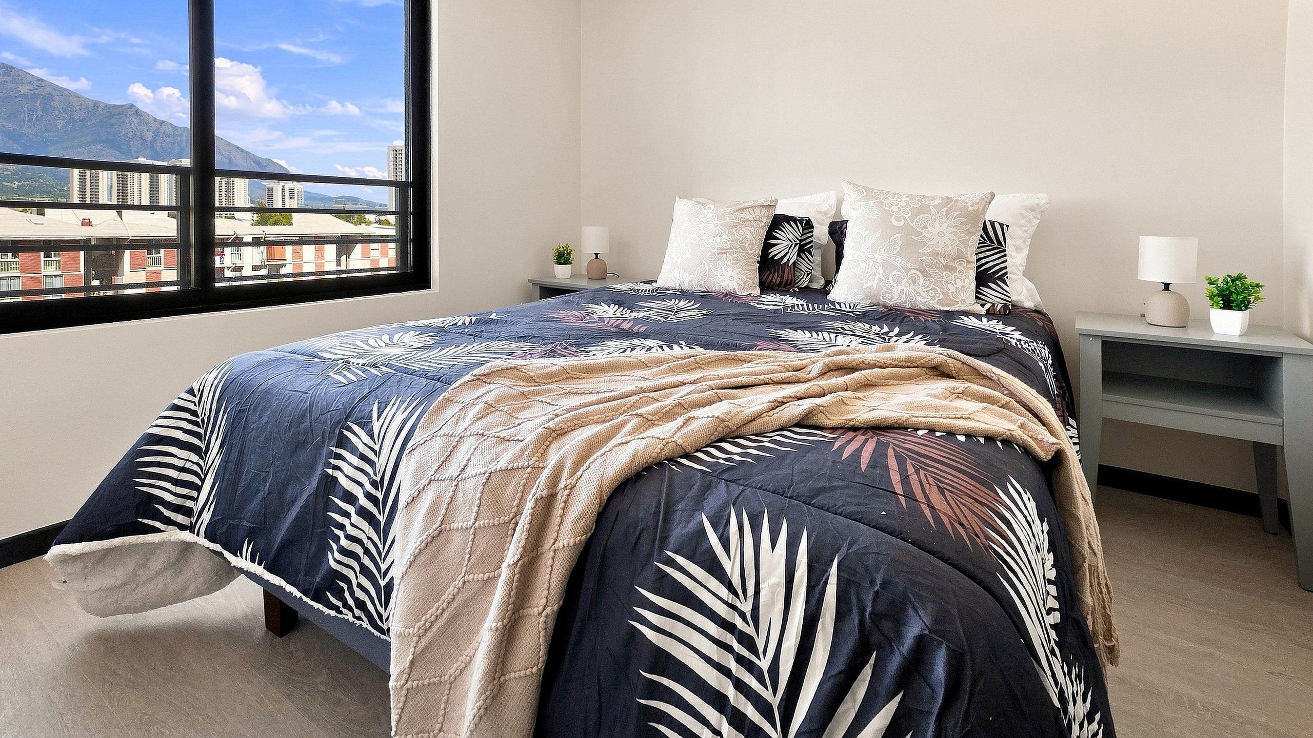 Un dormitorio con una cama, almohadas, una mesita de noche, una lámpara y una ventana con vistas a edificios y montañas.