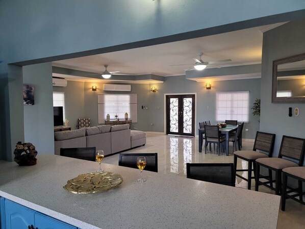 A modern kitchen with a central island, dining area with chairs, and a living room with a sofa and TV. - Villa Flores- New Modern Luxury Villa within walking distance from ANR Airport. (Bon Accord)