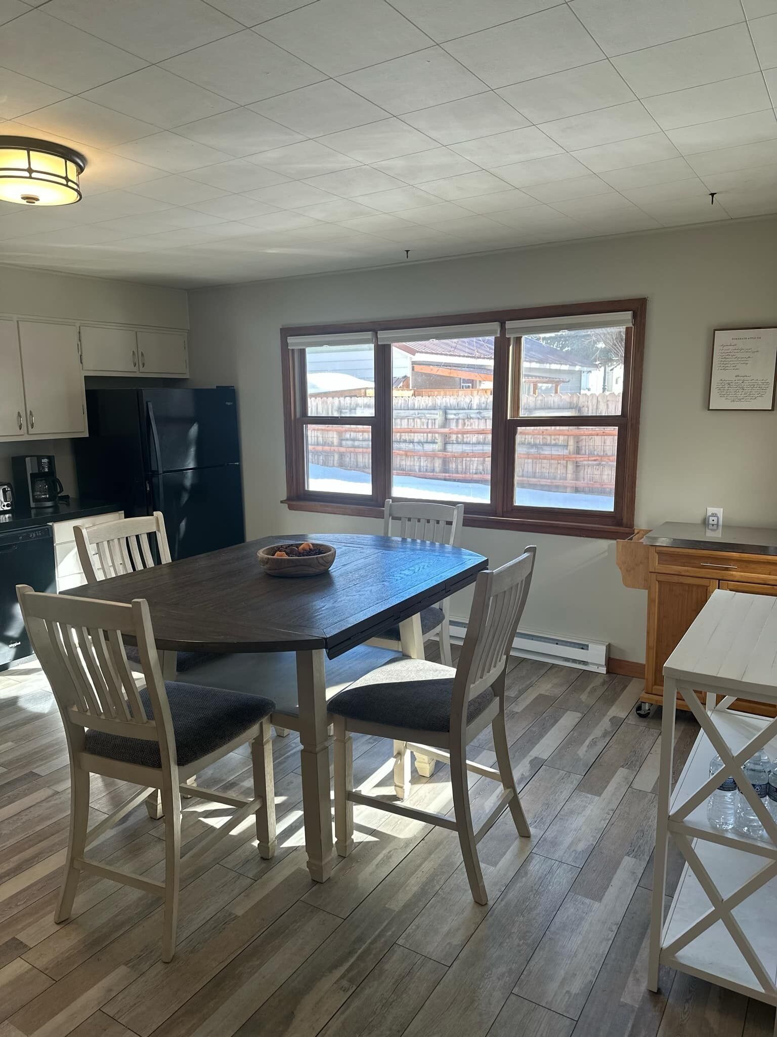 Un coin repas avec une table en bois foncé et des chaises blanches, un réfrigérateur noir et une fenêtre donnant sur un bâtiment traditionnel.