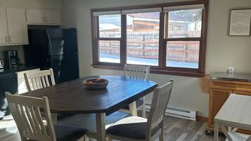 Un coin repas avec une table en bois foncé et des chaises blanches, un réfrigérateur noir et une fenêtre donnant sur un bâtiment traditionnel.