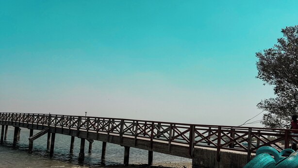 A beach with a wooden pier extending into the water, a sandy shore, and a large tire sculpture. - Hôtel Finio (Joal-Fadiout)