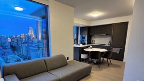 Un salon moderne avec un canapé gris, une table à manger avec des chaises et un coin cuisine doté de meubles sombres et d’un plan de travail blanc.