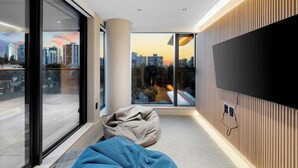 Un salon moderne avec un grand téléviseur, un fauteuil poire et une vue sur le paysage urbain au coucher du soleil.