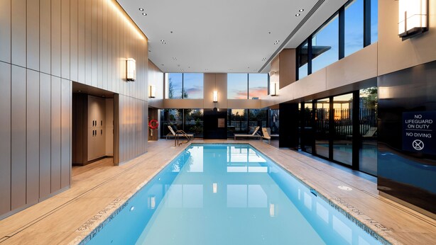 Une piscine intérieure moderne avec de grandes fenêtres offrant une vue sur le coucher du soleil, équipée de chaises longues et d’un panneau indiquant la présence d’un sauveteur.