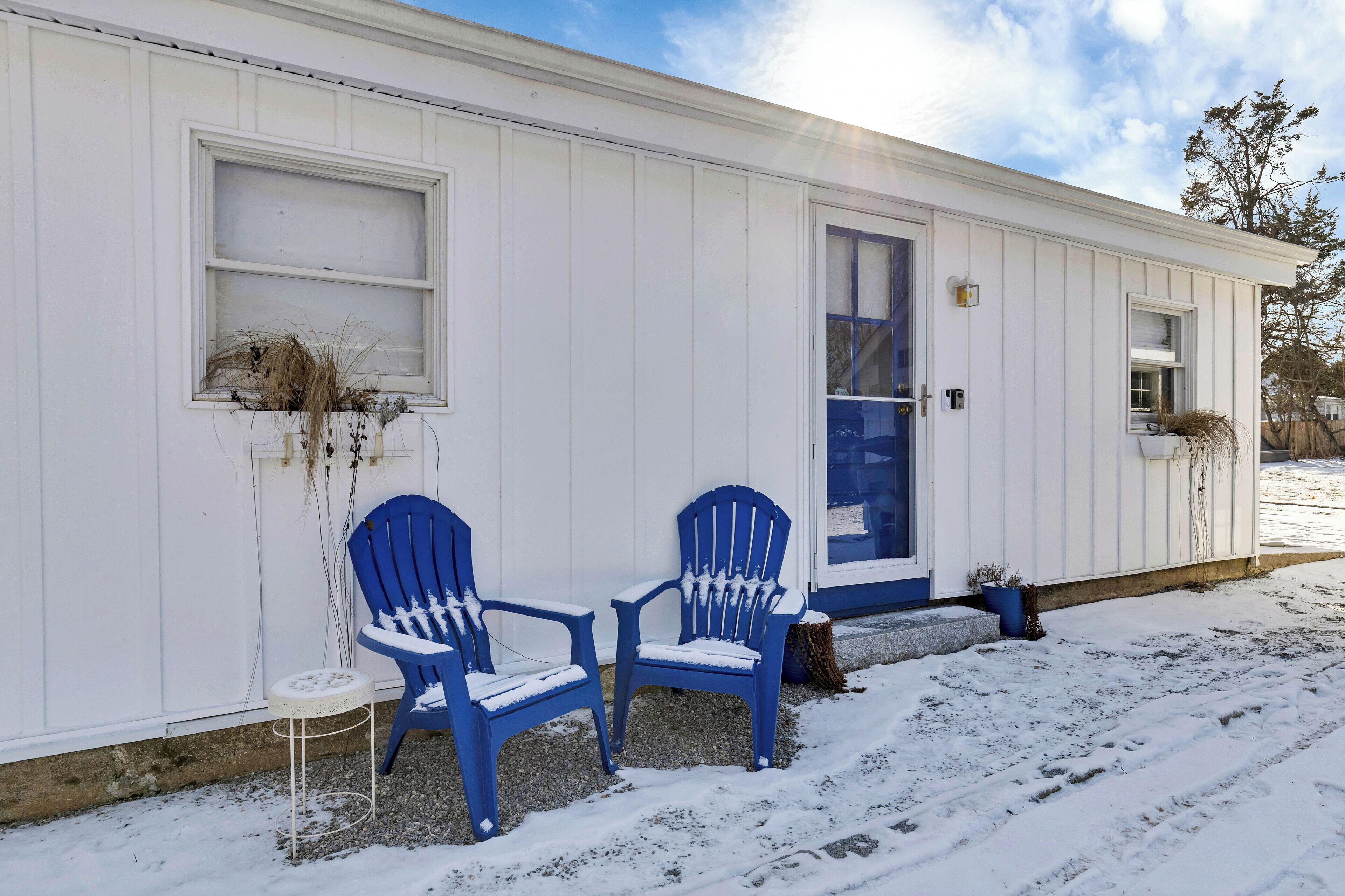 A white building with two blue Adirondack chairs outside, a small white table, and a window with a plant.