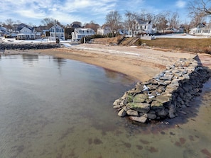 A waterfront property with a stone retaining wall, a sandy beach, and residential houses in the background.
